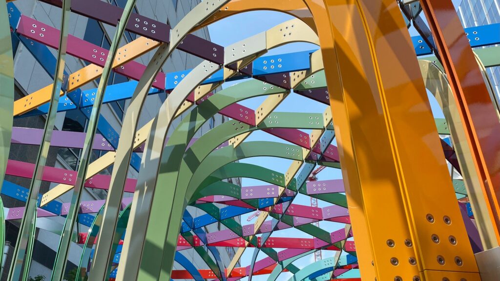 View of a metal sculpture from below with buildings and sky visible through it. The sculpture pieces are attached overhead like an arbor, and are multi-colored.