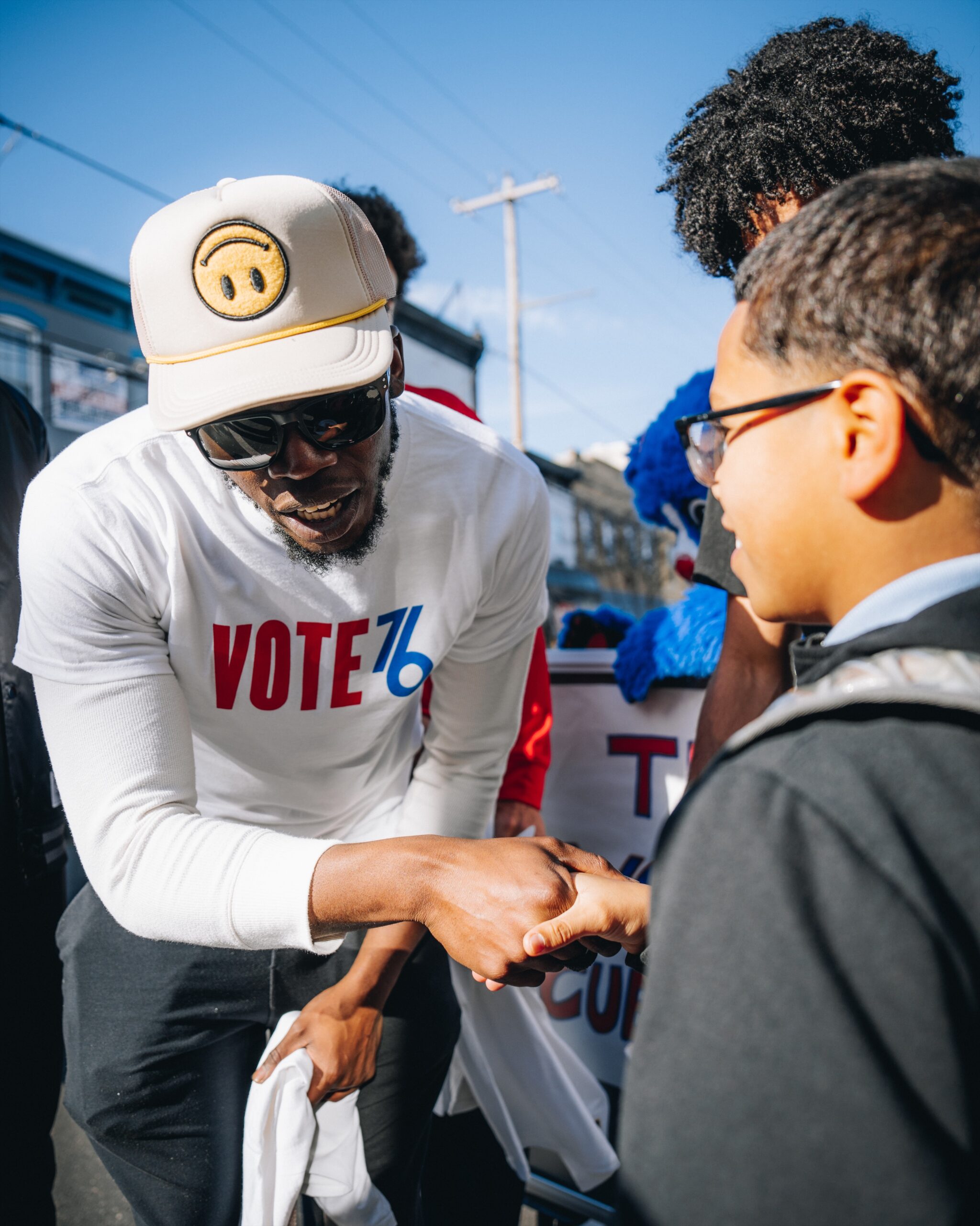 A man in a VOTE 76 shirt and hat shakes hands with a boy wearing glasses and a backpack at an outdoor event.