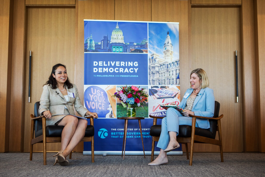 Two women sit in chairs smiling and talking in front of a Delivering Democracy banner. A bouquet of flowers sits between them. The setting appears to be a professional panel or discussion event.