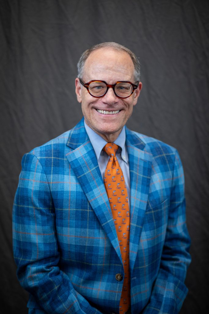 A person wearing a blue plaid blazer and an orange tie with a dolphin pattern smiles while wearing tortoiseshell glasses against a dark backdrop.